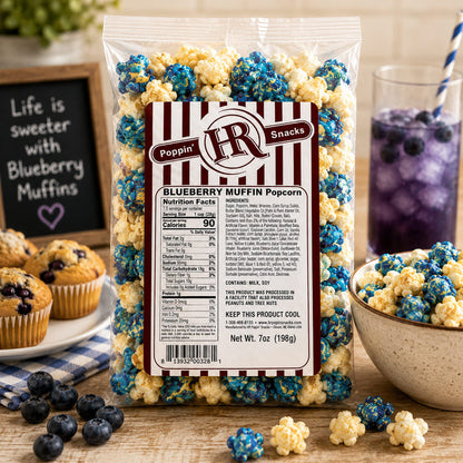 A 7 oz bag of Poppin' Snacks Blueberry Muffin Popcorn stands on a wooden table surrounded by blueberry muffins, a bowl of popcorn, blueberries, and a glass of purple drink. The bag has a clear plastic body with a large white and red striped label showing the brand 'Poppin' Snacks' and product details.