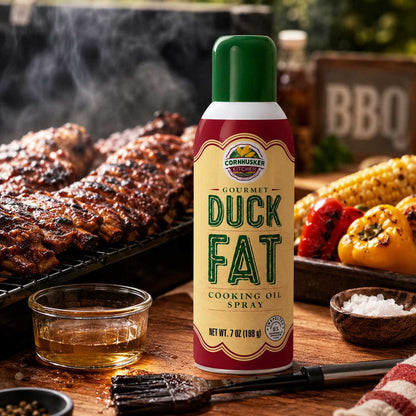 A can of Cornhusker Gourmet Duck Fat Cooking Oil Spray stands on a wooden table in a barbecue setting, with grilled meat, corn, peppers, salt, and a brush in the background.