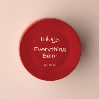 Red container of Trilogy Everything Balm on a beige background.