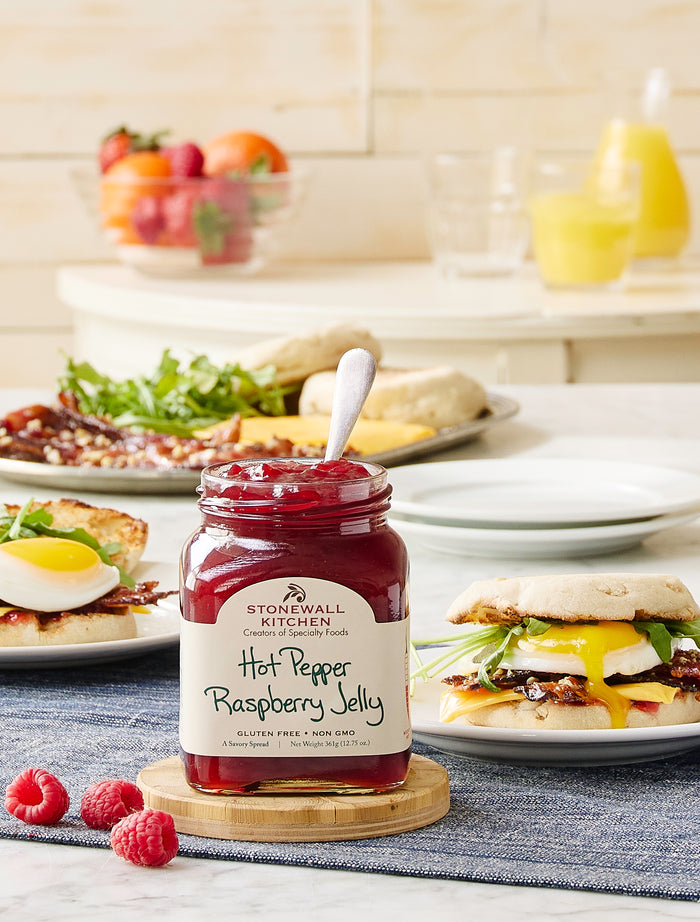 A jar of Stonewall Kitchen Hot Pepper Raspberry Jelly with a spoon in it, placed on a wooden coaster. In the background, there are breakfast foods including a fried egg on toast, an English muffin sandwich with bacon and greens, and a plate with bacon, greens, and biscuits. A bowl of fresh fruit and glasses of orange juice are also visible.