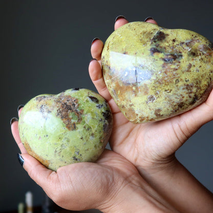 Two greenish-brown Opal stone hearts held in hands against a dark background